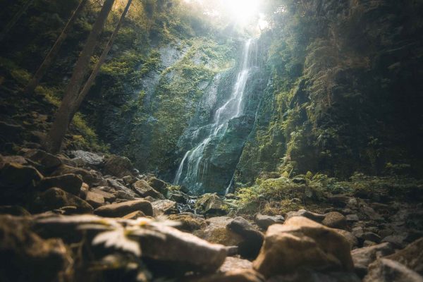 Burgbachwasserfall aus tiefer Froschperspektive über bemooste Felsbrocken, im unscharfen Vordergrund ein hell angeleuchtetes Blatt, oben bricht Sonnenlicht durch die Bäume.